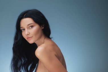 Koyu renk saçlı ve parlak tenli zarif bir kadın, doğal güzelliğini ön plana çıkaran yumuşak mavi bir arka plan karşısında yan profil çiziyor. Cilt bakımı, sağlık ve güzellik reklamcılığı kampanyaları için ideal.