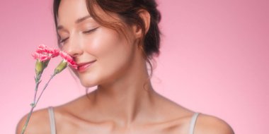 Genç bir kadının yumuşak ve romantik portresi. Gözleri kapalı gülümserken pembe karanfillerin kokusunun tadını çıkarıyor. Pastel pembe arka plan, doğal ışıltı ve dingin ifade.