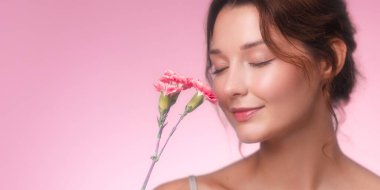 Stüdyo afişinde zarif, genç bir kadın var. Gözleri kapalı ve yumuşak bir gülümsemesi var. Nazikçe pembe karanfillerin kokusunun tadını çıkarıyor. Işıldayan cildi ve pastel pembe arka planı huzur verir.