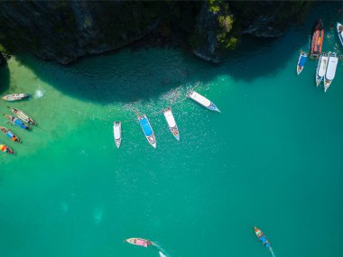 Tayland 'ın Ko Phi Phi kentindeki sarp kayalıkların yanında canlı turkuaz suya demirlemiş renkli teknelerin tepeden aşağı insansız hava aracı görüntüsü. Seyahat reklamları, egzotik varış yerleri, tekne gezileri ve ada turizmi için ideal..