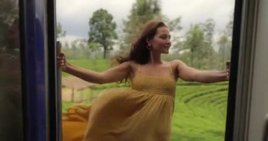 Sarı elbiseli bir kadın, Sri Lanka çay çiftliklerinin manzaralı güzelliğini kucaklayarak tren kapısından sarkıyor. Seyahat, macera ve yaşam tarzı promosyonları için idealdir.