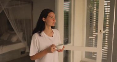 Gevşek beyaz gömlekli genç bir kadın sakin bir tropikal villada sıcak bir fincan kahvenin tadını çıkarıyor. Parlak bir kapının yanında duruyor, sabah ışığını ve sakin atmosferi kucaklıyor..