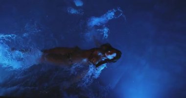 Göz kamaştırıcı bir kadın gece mavi bir havuzda zarif bir şekilde yüzer, yukarıdan süper yavaş çekimde yakalanır. Havanın perspektifi büyüleyici hareketi ve parlak suyu vurgular.. 