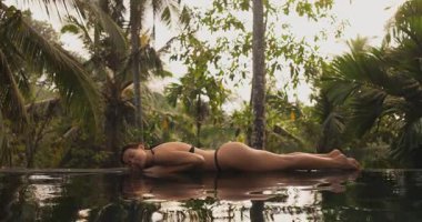 Göz kamaştırıcı bir bikini modeli, verimli tropikal yeşilliklerle çevrili sonsuz bir havuzun kenarında baştan çıkarıcı bir şekilde yatar. Ağır çekim sahnesi saf bir rahatlama ve şehvet anı yakalar.. 