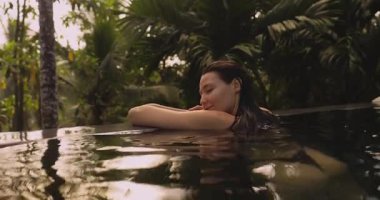 Bikinili sakin bir kadın sonsuz bir havuzun kenarına yaslanır ve ormanın sakin ortamının tadını çıkarırken gülümser. Bu sahne rahatlamayı, lüksü ve barışçıl bir tropikal kaçışı simgeliyor..