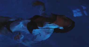 Siyah bikinili güzel bir kadın gece parlayan mavi suda huzur içinde yüzüyor. Ruhsal aydınlatma ve hafif dalgalanmalar büyüleyici ve rüya gibi sinematik bir atmosfer yaratır.. 