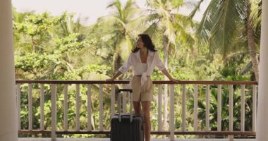 Zarif bir kadın bavuluyla birlikte tropik bir tatil köyünün balkonunda duruyor ve otelden ayrılmadan önce bereketli palmiye ağacı manzarasına son bir kez bakıyor.. 