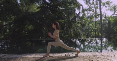 Genç bir kadın, zengin tropikal yeşilliklerle çevrili sonsuz bir havuzun yakınındaki yoga seansı sırasında Warrior pozu veriyor. Doğada dengeyi, gücü ve farkındalığı kucaklıyor.. 