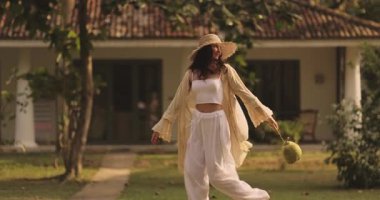 Hasır şapkalı ve akıcı kıyafetli şık bir kadın elinde taze hindistan ceviziyle tropikal bir otelde neşeyle yürüyor. Tatil, rahatlama ve doğa için mükemmel bir yaz sahnesi.. 