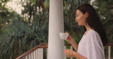 Aşırı büyük beyaz gömlekli sakin bir kadın, bereketli tropikal yeşilliklerle çevrili manzaralı bir balkonda kahvesini yudumluyor. Huzurlu bir sabah atmosferini kucaklıyor, rahatlığın ve farkındalığın tadını çıkarıyor..