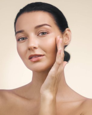 Işık saçan, nemli ciltli ve yüzüne nazikçe dokunan makyajlı genç bir kadının stüdyo yakın çekimi. Cilt bakımı reklamları, güzellik ürünü görselleri ve kişisel bakım promosyonları için ideal.