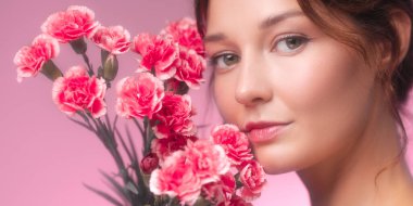 Yüzüne yakın pembe karanfiller tutan güzel bir kadının geniş formatlı stüdyo görüntüsü. Romantik ve zarif bir görünüş kozmetik, çiçek ve sağlık promosyonları için mükemmel..