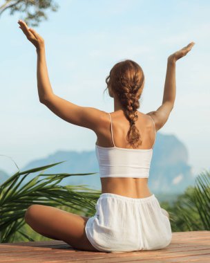 Genç bir kadın kollarını kaldırarak çapraz bacaklı bir pozisyonda meditasyon yapıyor ve tropik bir dağ manzarasına bakıyor. Sağlık, ruhsal inzivaya çekilme ve doğadan esinlenilmiş promosyon için idealdir..