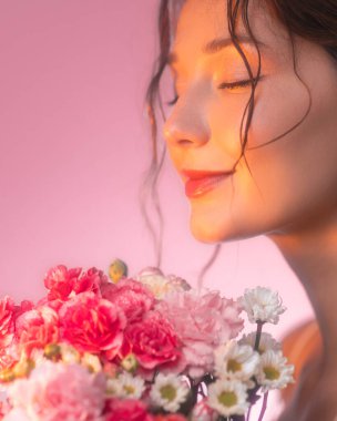 Işıl ışıl cildi ve gözleri kapalı pembe ve beyaz çiçekleri koklayan bir kadının yumuşak odaklı güzel portresi. Altın gün batımı ışığı rüya gibi ve sakin bir atmosfer ekler..