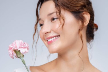 Tarafsız bir arka planda soluk pembe bir çiçek tutan gülümseyen beyaz kadın. Temiz güzellik kavramı cilt bakımı, kozmetik endüstrisi, sağlık ve kadın markalaşması için idealdir..