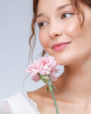 Yüzünün yanında pembe karanfil tutan gülümseyen beyaz bir kadının taze ve genç portresi, doğal güzellik kavramı, cilt bakımı, çiçek yaşam tarzı ve kozmetik reklam tasvirleri..