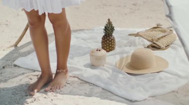 Sahil kenarında hasır şapkası, hindistan cevizi içeceği, ananas ve beyaz kumaşla örülmüş bir çanta. Beyaz elbiseli çıplak ayaklı bir kadın bu sakin tropikal sahneyi tamamlıyor. Yaz ya da yaşam tarzı için mükemmel