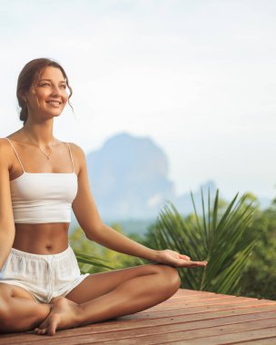 Beyaz spor giysili genç bir kadın Tayland, Krabi 'de yemyeşil ve kireçtaşı dağlarıyla çevrili ahşap bir güvertede keyifli bir meditasyon pozisyonunda oturuyor. Sağlık ve yaşam tarzı içeriği için ideal.