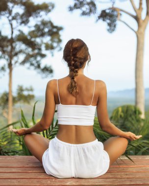 Arkadan görülen beyaz kadın, yemyeşil bir ormanın ortasında tahta bir güvertede meditasyon yapıyor. Sağlık reklamları, doğa geri çekilmeleri ve yaşam tarzı markalaştırma malzemeleri için ideal..