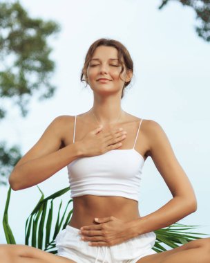 Beyaz kadın meditasyonu tropikal ortamda göğüs ve karın bölgelerine yerleştirilmiş elleri ve gözleri kapalı yapar. Topraklamayı, iyileştirmeyi ve farkındalığı sembolize eder. Refah içeriği için ideal.