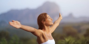 Beyaz kadın gözleri kapalı ve kolları açık gülümser, tropik bir dağ manzarasında özgür hisseder. Sağlık, özgürlük, yaşam tarzı ve seyahat içeriği için mükemmel..