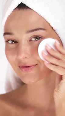 Beyaz bir havlu giyerken yüzüne pamuk pedi süren beyaz tenli bir kadın cilt bakımı, yüz bakımı ve reklam kullanımı için modern güzellik kavramlarını vurguluyor..