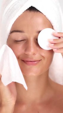 Havlu giymiş beyaz bir kadın pamuk ped ve temizleyici mendil kullanarak makyajı nazikçe temizliyor. Güzellik markaları, cilt reklamları, makyaj temizleyiciler ve sağlıklı yaşam tarzı görselleri için mükemmel..