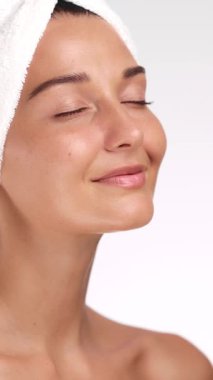 Beyaz tenli ve kafasında havlu olan beyaz bir kadın yüzüne hafifçe dokunuyor, cildini, sağlığını ve güzellik anlayışını yüksek kaliteli kaplıca ortamında temsil ediyor..