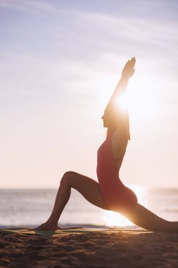 Pembe aktif giysili bir kadın silueti gün batımında plajda yoga esnemesi yapıyor. Sakin okyanus arkaplanı ve batan güneşin ılık parıltısı sakin, düşünceli bir atmosfer yaratır.