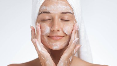 Gözleri kapalı sakin bir kadın yüzünü köpüklü bir temizleyiciyle yıkamaktan hoşlanır. Bir havluya sarılmış, kendine bakımı, cilt bakımını ve sağlığını kucaklıyor, sağlıklı, parlayan bir cildi teşvik ediyor.. 