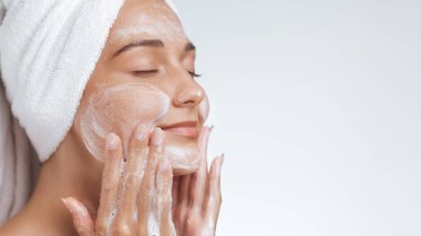 Kafasına havlu sarılmış rahat bir kadın yüzünü köpüklü sabunla nazikçe temizliyor ve cilt bakımının tadını çıkarıyor. Güzellik, özgüven ve sağlıklı cilt temaları için mükemmel..