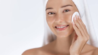 Parlak bir gülümsemesi olan genç bir kadın havluya sarılmış halde yüzünü pamukla nazikçe temizliyor. Güzellik, cilt bakımı ve kişisel bakım için mükemmel bir kavram.. 