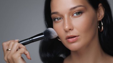 Göz kamaştırıcı bir kadının, kusursuz cildini ve güzellik rutinini vurgulayarak yumuşak bir fırçayla makyaj yaptığı yakın plan. Güzellik, cilt bakımı ve kozmetik ürünler için ideal promosyonlar veya reklamlar.