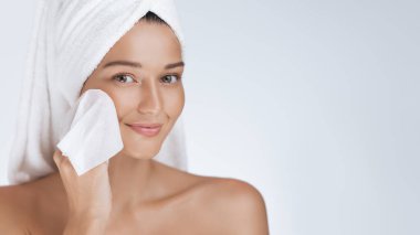 Kafasında havlu olan güzel genç bir kadın makyaj temizleyicisiyle yüzünü nazikçe temizliyor. Taze, ışıl ışıl cilt için cilt bakımı. Güzellik ve sağlık promosyonları için mükemmel..
