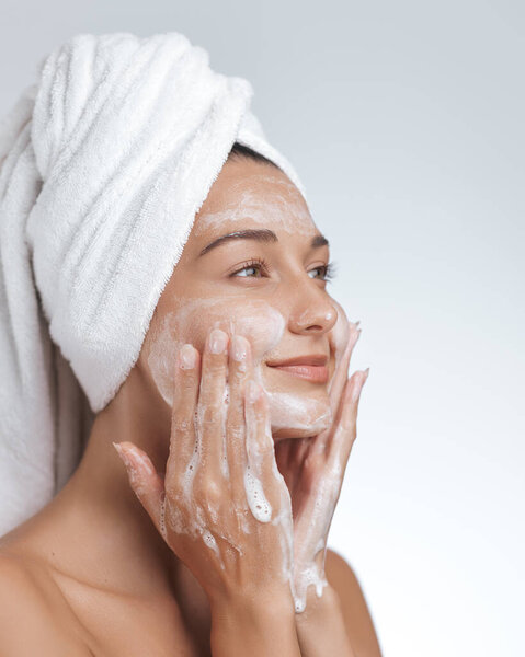 Молодая женщина нежно умывается пенным очистителем, надевая полотенце на голову, захватывая освежающий ритуал очищения кожи. Идеально подходит для продвижения продуктов по уходу за кожей, здоровья и красоты