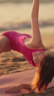 Dar pembe elbiseli güzel kadın gün batımında plajda zarif yoga pozuyla esniyor. Kavramsal ve duygusal sağlık videosu, fitness, yaşam tarzı ve tanıtım amaçları için ideal.