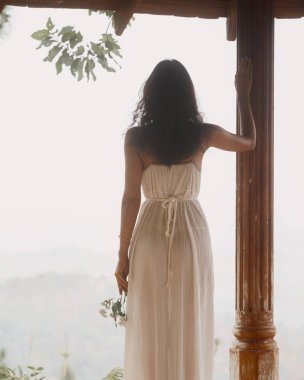 Pilili beyaz elbiseli zarif beyaz bir kadın elinde çiçekler taşralı bir sütunun yanındaki mesafeye bakıyor. Romantik, spa, moda ve huzurlu yaşam tarzı görselleri ve reklamları için mükemmel..