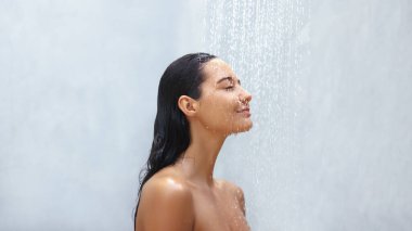 Sakin bir kadın, bir duş sırasında hafif bir su akıntısının altında durur, gözleri gevşemek için kapalıdır. Bu sakin kişisel bakım anı güzellik, güzellik ve cilt bakımı ile ilgili konular için mükemmeldir.