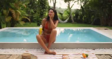 Canlı bikinili rahat bir kadın köpüklü bir havuzun kenarında oturmuş, elinde canlandırıcı bir şişe meyve suyu tutuyor. Tropikal yeşilliklerle çevrili bu sahne boş zamanları, refahı ve tatil temalarını vurguluyor.. 