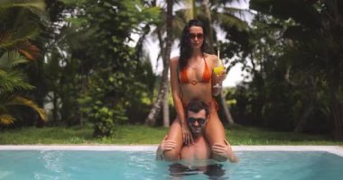 Tropikal bir yüzme havuzunda eğlenen oyuncu bir çift. İçkili bikinili bir kadın yemyeşil bir ortamda erkeğin omuzlarında oturuyor. Tatil ve yaşam tarzı kavramları için mükemmel.. 