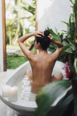 Saçı kabarık sakin bir kadın tropikal yapraklar ve mumlarla çevrili açık havada bir küvette rahatlıyor. Spa, sağlık, cilt bakımı, doğal güzellik ve lüks yaşam tarzı kampanyaları için mükemmel..