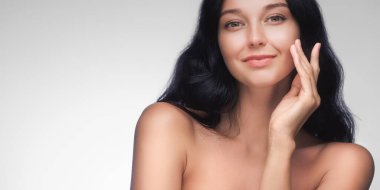 Koyu renk saçlı ve parlak tenli zarif bir kadın yüzüne dokunurken hafifçe gülümseyip doğal güzelliğini ön plana çıkarıyor. Sağlık, cilt bakımı ve güzellik pazarlama kampanyaları için ideal.