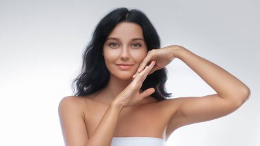 Koyu renk saçlı ve açık tenli genç bir kadının zarif portresi. Minimalist beyaz bir stüdyoda elleri yüzüne yakın poz veriyor. Güzellik, cilt bakımı ve sağlık kampanyaları için ideal..