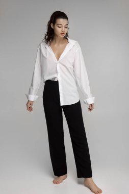 Beyaz gömlekli, siyah pantolonlu, rahat bir ifadeyle yalınayak poz veren genç bir kadın. Moda yazıları, minimalist yaşam tarzı ve günlük zarafet temaları için mükemmel.. 