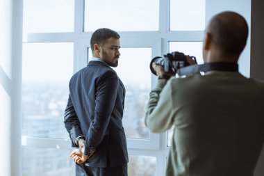 Profesyonel bir fotoğrafçı büyük ofis pencereleri önünde poz veren şık bir işadamını iş başyazıları, marka çekimleri ve idari profil içeriği için mükemmel bir arka planla yakalıyor..