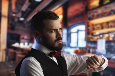 Sakallı ve resmi kıyafetli kendine güvenen genç adam saatini gece hayatı, dakiklik, bekleme ve modern erkeklik tanıtımı için mükemmel bir şekilde aydınlatılmış bir barda kontrol ediyor..
