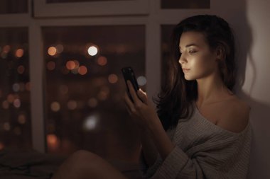 Geceleri pencerenin kenarında oturan, akıllı telefon ekranı tarafından aydınlatılmış güzel genç bir kadın. Arka plandaki sıcak şehir ışıkları rahat ve modern bir ortam yaratıyor. Teknoloji ve yaşam tarzı için mükemmel.