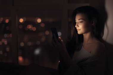 Geceleri pencerenin yanında oturan şık genç bir kadın arka planda parlayan şehir ışıklarıyla akıllı telefonunu kullanıyor. Yaşam tarzı, teknoloji ve modern yaşam kavramları için mükemmel.. 