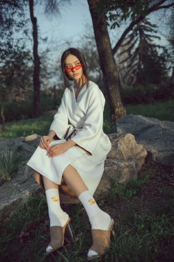 Beyaz kimonolu modaya uygun Asyalı kadın, doğal park ortamında kayaların üzerinde oturuyor. Günbatımında kavramsal moda portresi, editör, reklam veya tanıtım kampanyası görselleri için mükemmel.