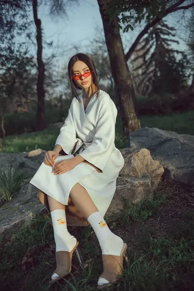 Beyaz kimonolu modaya uygun Asyalı kadın, doğal park ortamında kayaların üzerinde oturuyor. Günbatımında kavramsal moda portresi, editör, reklam veya tanıtım kampanyası görselleri için mükemmel.